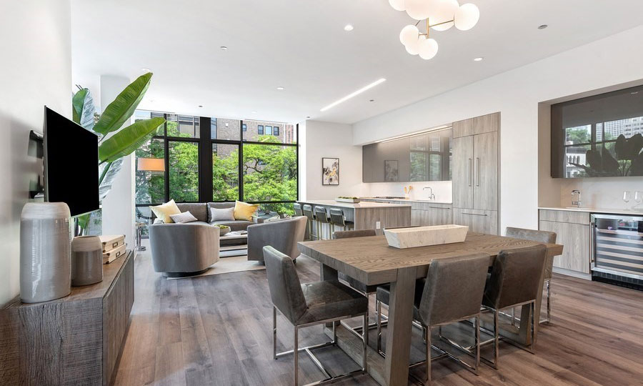 A dining set is in the foreground, a living room set behind it along floor-to-ceiling windows. In the back right is the kitchen with a counter-top and stools.