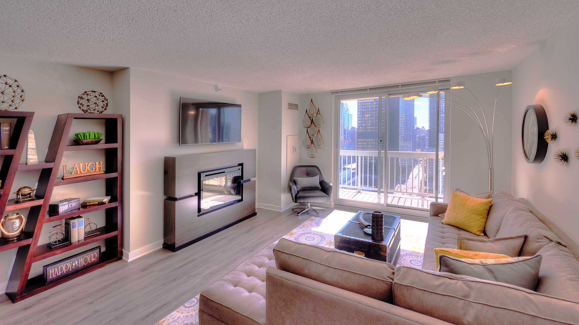 Looking into a living room with a sliding glass window leading to a balcony on the far end. A sitting area is on the right with a large couch and table, across is fireplace and mounted television..