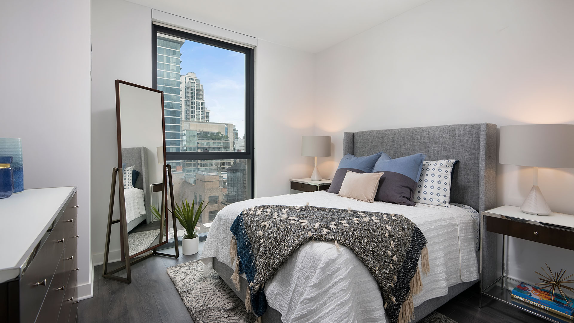 A bedroom in a Hubbard221 residence. The bed in on the right with end tables on both sides. There is a tall window past the bed with a standing mirror beside it. There is a dresser on the left wall.